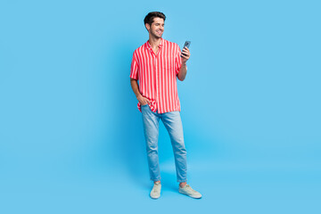 Fototapeta na wymiar Full body photo of clever cheerful guy wear stylish shirt jeans trousers look at smartphone read email isolated on blue color background