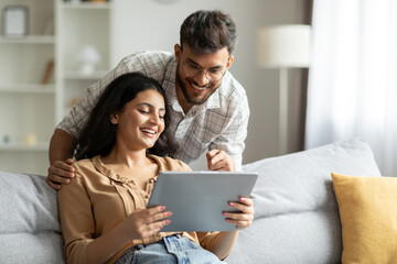 Happy indian spouses using digital tablet, surfing internet or shopping online while resting on sofa at home, free space. Hindu man and woman relaxing with modern gadget