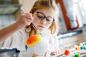 Little girl painting globe or ball with colors. School child making earth globe for school project. Happy kid with eyeglasses holding brush.