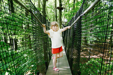 Cute little preschool girl walking on high tree-canopy trail with wooden walkway and ropeways on...
