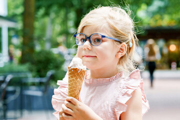 Little preschool girl with glasses eating ice cream in waffle cone on sunny summer day. Happy...