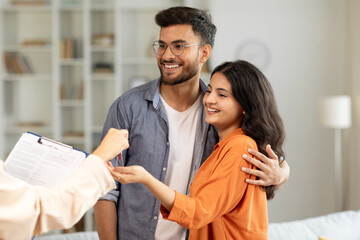 Real estate agent giving keys to young indian buyers, happy woman taking key of future new...