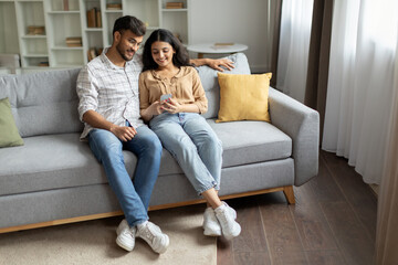 Obraz premium Indian couple using cellphone together, sitting in casual home interior on sofa, surfing internet or chatting with friends. Modern, digitally connected lifestyle