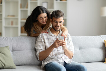 Young hindu couple sharing social media on cellphone, happy indian spouses sitting on sofa, using smartphone gadget at home interior, woman embracing husband from back