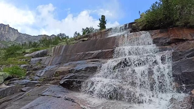 A stormy stream cascades down from a high rocky waterfall on a sunny summer day. Marble Waterfall, Natural Prak Ergaki, Krasnoyarsk Territory, Siberia, Russia.
