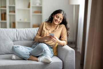Smiling indian lady with smartphone resting on couch at home, woman texting with friends, surfing...