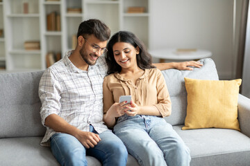 Happy indian spouses using mobile phone while relaxing on sofa, scrolling social media app or...