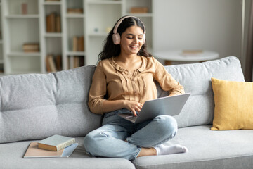 Obraz premium Indian lady wearing headphones and using laptop, enjoying digital leisure and e-learning, sitting on sofa at home interior, copy space