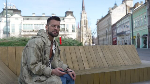 Young hipster millennial man sitting on the bench on the street waiting for woman he met online on online dating site or social network. Nervous male expect female that they organized blind date