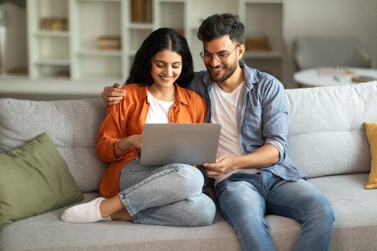Loving indian spouses using laptop together, sitting on sofa in cozy home interior, man hugging his wife and looking at computer screen, choosing movie