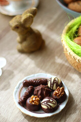 Easter eggs in the basket, bowl of cookies, chocolate pralines, Easter bunny figurine, cups of tea, glasses of juice, flowers and lit candles on the table. Selective focus.