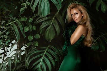 Elegant woman in a green evening dress poses sensually against the background of Monstera plants