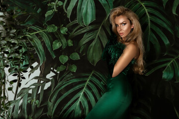 Fototapeta na wymiar Elegant woman in a green evening dress poses sensually against the background of Monstera plants
