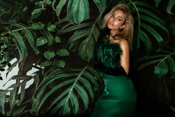 Elegant woman in a green evening dress poses sensually against the background of Monstera plants