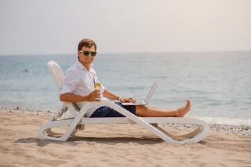 Young man in sunglasses and white shirt is lying on a beach chair on the seaside holding a cocktail and a laptop.