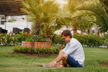 Young man in sunglasses and white shirt is lying on grass  and working in a tropical location