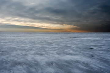 A frozen sea covered with dark ice and a thin layer of snow. Cloudy sky and orange sunlight at the...
