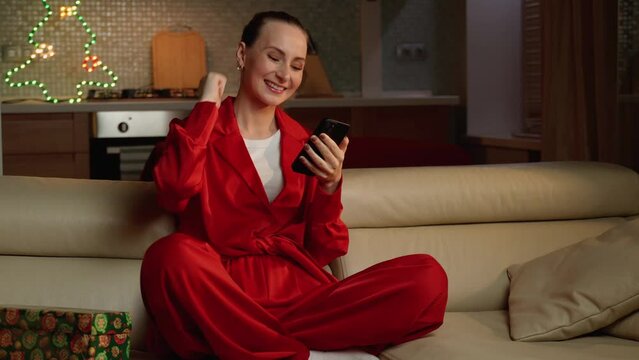 A smiling woman in cozy red pajamas is enjoying some leisure time on her couch with a smartphone in her hand, in a warmly lit living room.
