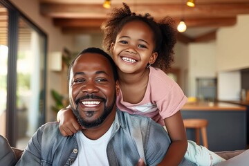 An African American dad is pictured with his daughter hugging him from behind, both smiling