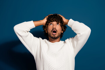 Portrait of young amazed indian funny man wearing white pullover touch head look above head isolated on dark blue color background