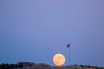 This captivating image captures the full moon rising majestically behind a Turkish flag atop an...