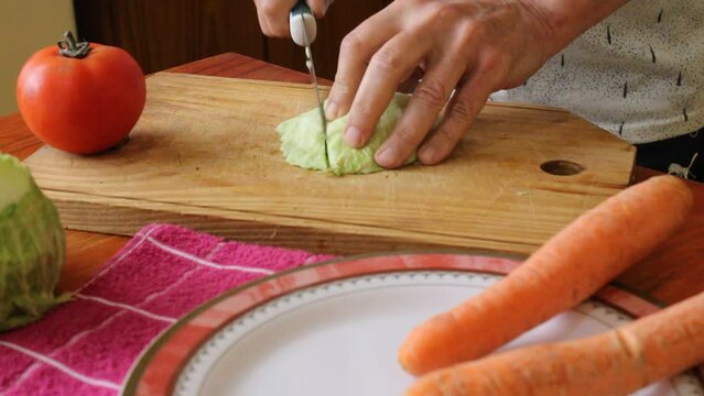 Comida t&iacute;pica vegana sudamericana Argentina, el hombre prepara un almuerzo vegetariano en la mesa de madera de casa, corta con el chuchillo repollo  tomate y zanahorias en un plato de cocina saludable