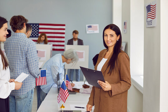 Portrait of American female poll worker. Happy beautiful young brunette woman in suit standing at US ballot station, holding clipboard, looking at camera and smiling. Voting, election concept