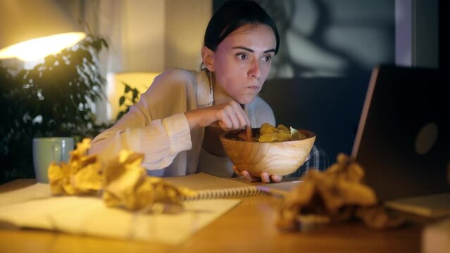 Happy positive woman with chips laughing while watching comedy movie show serials instead of work or education at home workplace Tired female relaxed after hard work day with snack at night indoors