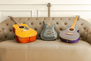 Three guitars set on a brown leather sofa in the living room. A black electric guitar, a brown...