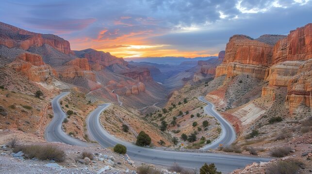 a winding road in the middle of a mountain range with a sunset in the background and clouds in the sky.
