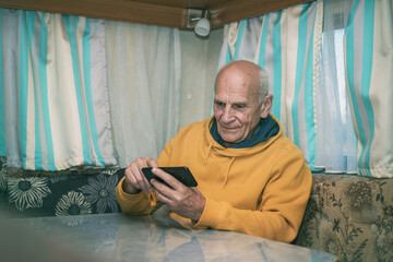 Older man sitting at desk using and checking smartphone in camping rv in style of dark yellow and...