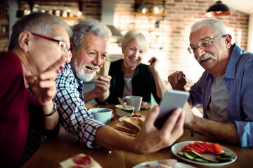 Senior friends laughing and sharing photos on smartphone at breakfast