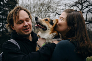 A young man and woman are smiling and hugging with their cute Welsh corgi puppy in the park. Dog...
