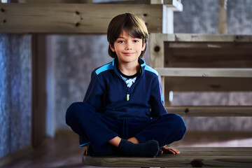 Caucasian male child of seven years old sits cross-legged on step of wooden staircase at home.