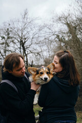 A young man and woman are smiling and hugging with their cute Welsh corgi puppy in the park. Dog with happy expression on its face. Life style portrait