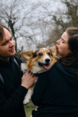 A young man and woman are smiling and hugging with their cute Welsh corgi puppy in the park. Dog with happy expression on its face. Life style portrait