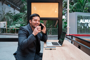 Cheerful latin man talking on his smart phone while sitting in a bar drinking coffee and working on his laptop.