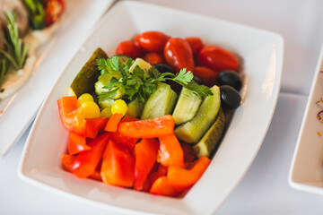 Deep white plate with various vegetables cucumber, cherry, pepper stands on table in restaurant. 