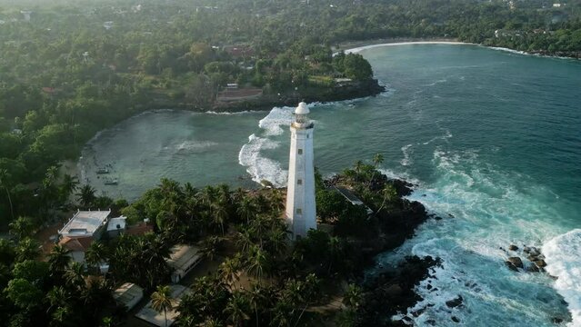 Drone lighthouse on rocky point at Dondra Head, Sri Lanka. Waves crash against cliffs, surrounded by greenery. Historical beacon stands tall, guiding sailors. Tranquil bay viewed from above.