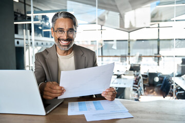 Happy mature professional business man lawyer holding documents in office. Middle aged male ceo executive, entrepreneur or bank manager sitting at desk in corporate office. Portrait, copy space.