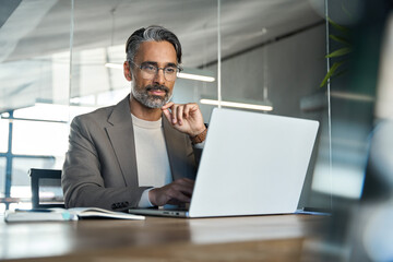 Busy middle aged Latin business man executive ceo wearing suit and glasses sitting at desk using...