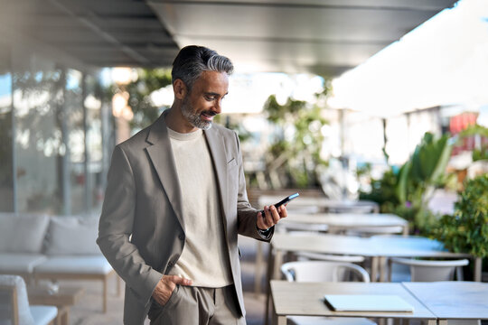 Happy smiling middle aged business man, rich mature professional businessman entrepreneur standing in outdoor cafe holding smartphone using mobile cell phone digital tech. Authentic candid photo.