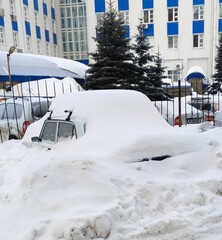 There is a car near the fence, covered with a thick layer of snow against the background of a building and fir trees