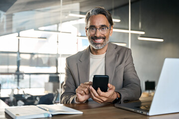 Fototapeta na wymiar Happy mature 45 years old business man executive sitting at desk using mobile phone. Smiling middle aged businessman looking at camera working on computer technology holding smartphone. Portrait.