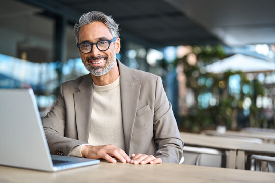 Happy confident mature professional business man hybrid working looking at camera. Smiling middle aged businessman wearing glasses sitting at outdoor office table using laptop. Portrait. Copy space.