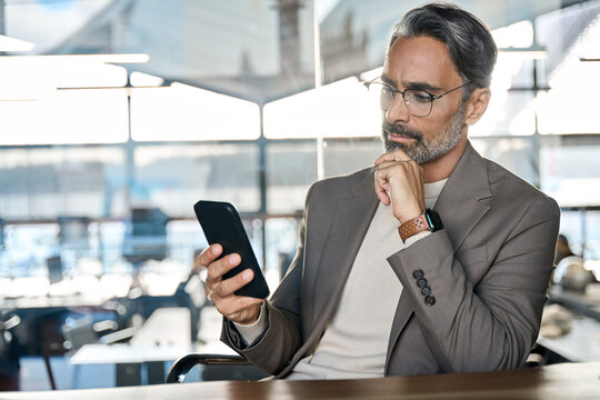 Busy mature business man investor thinking looking at mobile cell phone in office. Concerned or doubtful middle aged businessman executive using smartphone analyzing financial investment risks.
