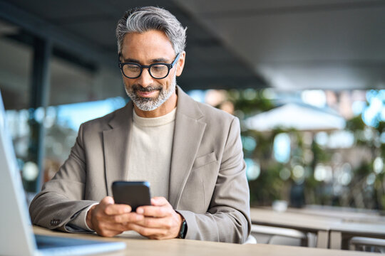 Happy middle aged business man using mobile phone sitting outside office. Stylish older busy businessman investor wearing glasses holding smartphone looking at cellphone doing financial payments.