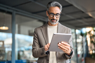 Busy mature older business man executive standing in office using digital tablet. Middle aged...