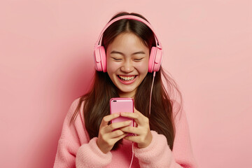 Happy cheerful joyful positive young Asian woman on pastel studio background uses mobile phone and noise cancellation headphones head set, listens to music, has good time, enjoys her favorite tracks - obrazy, fototapety, plakaty