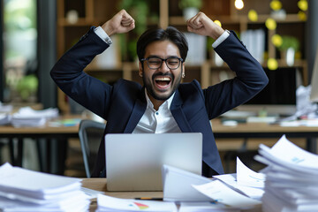 Happy excited young Indian business man accountant standing at the desk working on laptop computer with a pile of documents on table in office and making yes gesture rejoicing in successful job.